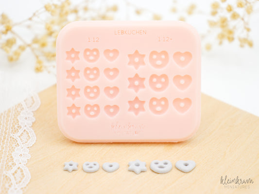 A pink silicone mould with indented shapes for making miniature soft gingerbread: a star, a pretzel, and a heart in small and large sizes. Three gray clay pieces for each size shaped like a star, a pretzel, and a heart, displayed below the silicone mould.