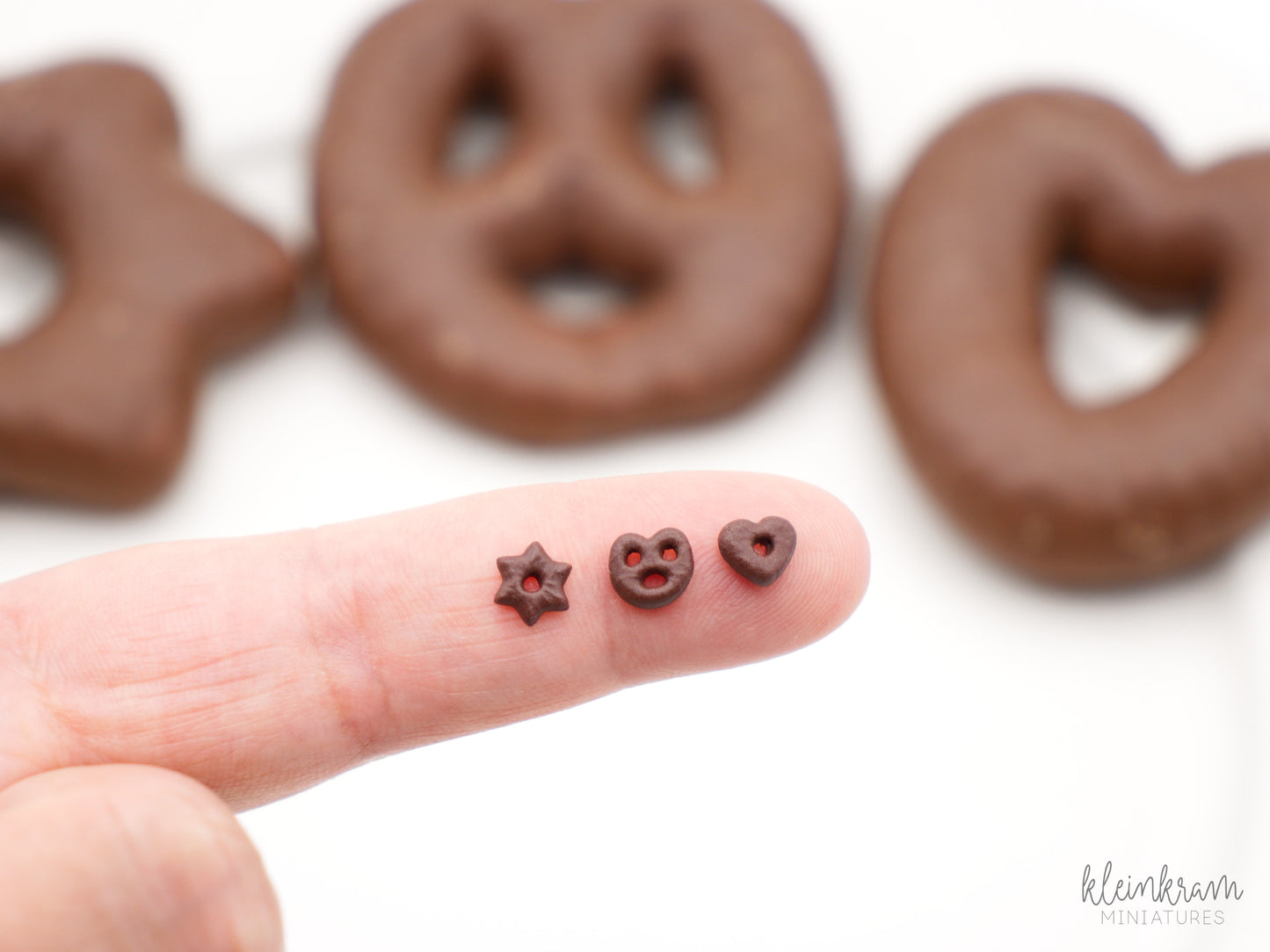 Three miniature soft gingerbread, lebkuchen in 1:12 scale (one inch scale) in the shapes of a star, a pretzel and a heart on a finger. The real life sized version are in the background.